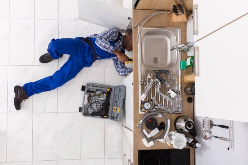 A plumber laying on his back underneath a kitchen sink performing a repair with a tool case laying next to him