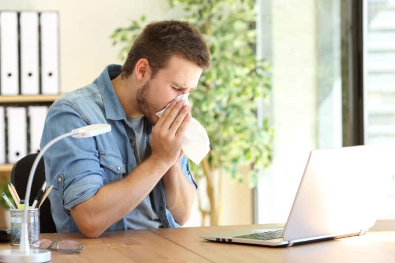 A man on his laptop being affected by allergies and sneezing into a tissue